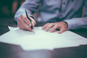 A man in a blue button down shirt signing papers on a wood table with a pen.